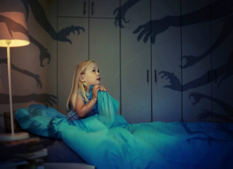 A young girl is sitting in bed, holding a blanket up to her face, looking to the side with a concerned expression. Shadowy hand figures are projected on the walls around her, creating a spooky atmosphere as she deals with nightmares. The room is dimly lit by a bedside lamp.