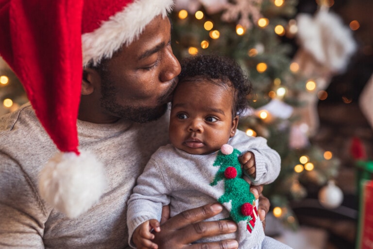 A man wearing a Santa hat holds and kisses a baby dressed in a gray sweater with a Christmas tree design, capturing photos of baby's first Christmas. They are in front of a decorated Christmas tree with lights.