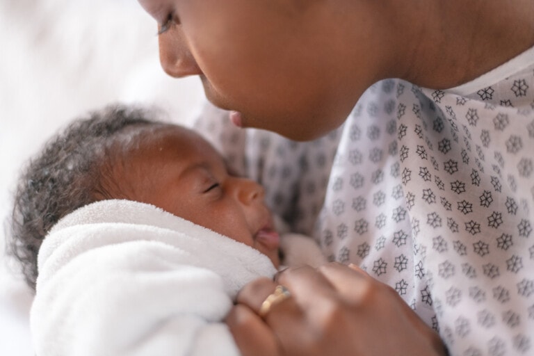 A person wearing a patterned garment holds a sleeping baby wrapped in a white blanket. The person's face is close to the baby, and they appear to be looking down at the infant, much like one might gaze at someone peacefully resting under weighted blankets.