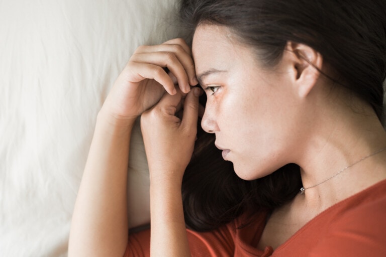 A woman wearing an orange top lies on her side on a white surface. Resting her head on her hand, she appears to be deep in thought, possibly reflecting on PMADs and their impact.