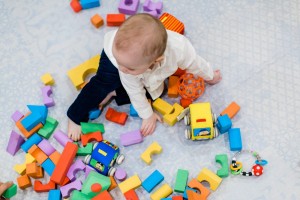 A toddler wearing a white shirt and black pants is sitting on a light-colored floor, surrounded by colorful baby items like toys and building blocks. The child is reaching for a purple block while other toys, such as a yellow toy car and a rattle, are scattered around.