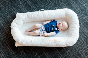 A baby lies in a white cushioned DockATot baby lounger placed on a geometric-patterned dark gray floor. The baby is dressed in a blue shirt with the number "02" and plaid shorts, looking up with legs extended.