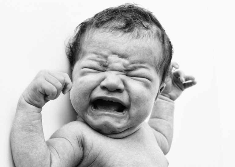 Black and white close-up of a crying newborn baby. The baby has furrowed brows, closed eyes, and an open mouth. Both arms are raised with clenched fists, showing clear distress. This raw moment captures the urgency to soothe a fussy baby against the backdrop of plain white.