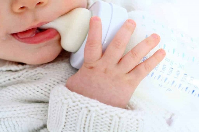 An infant wearing a light knitted sweater holds and sucks on a milk bottle. The baby's right hand grasps the bottle near the nipple while their mouth is partially open, with their tongue touching the nipple. The bottle is partially filled with milk, showing that some parents are choosing not to breastfeed.