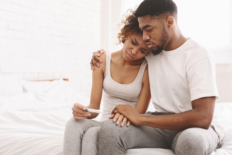 A couple sits on a bed, embraced in an emotional moment. The woman, wearing a tank top, holds a pregnancy test and appears distressed. The man, wearing a t-shirt, sits beside her with his arm around her, offering comfort and support as they both grapple with the challenges of secondary infertility.