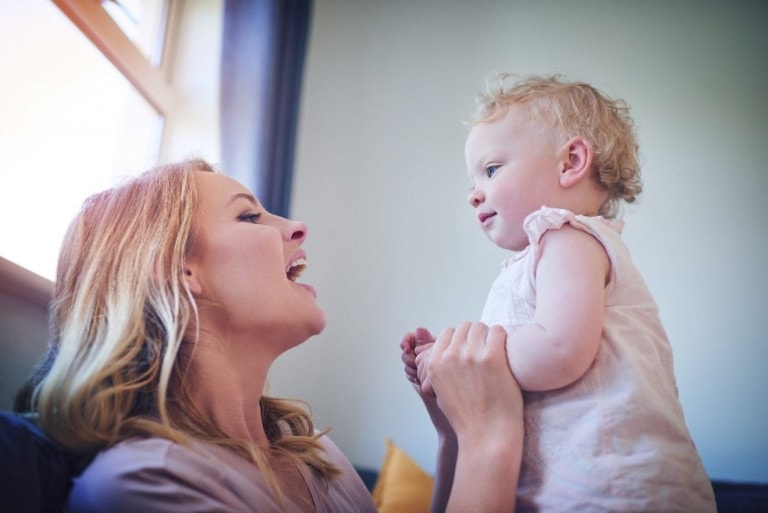A woman with shoulder-length blonde hair is holding a baby. The woman is smiling and looking up at the baby, who is standing and looking back at her. They are indoors, with a window in the background letting in natural light. Scenes like these often capture the precious moments before babies start talking.