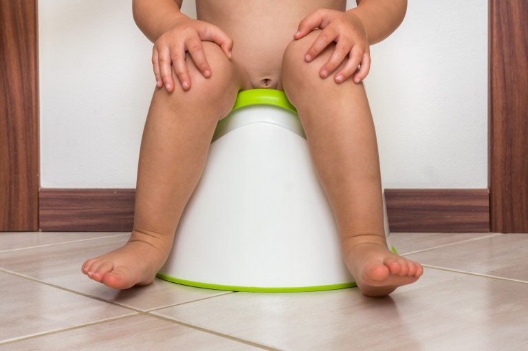 A toddler, possibly experiencing potty training too early, sits on a white potty with green accents. The child's legs and feet are visible, with hands resting on their thighs. The scene is set indoors on a tiled floor, against the backdrop of a wall and a wooden door frame.