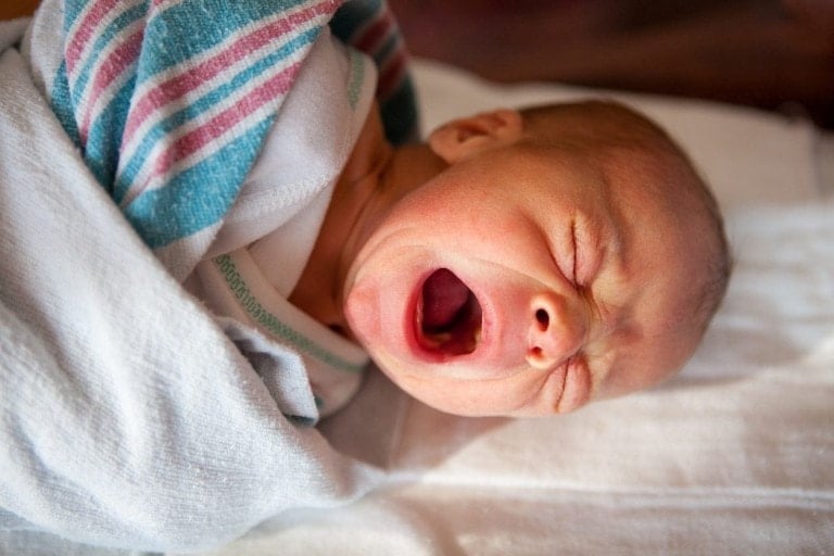 A newborn baby wrapped in a white blanket with teal and pink stripes is lying down and crying, possibly due to a tongue tie. The baby's eyes are closed, and their mouth is wide open. The background is out of focus.