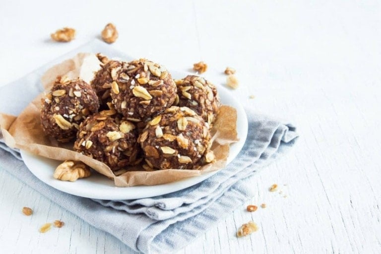 A white plate on a light wooden table holds five energy balls made from nuts and oats, perfect lactation snacks, lined with brown parchment paper. A few loose nuts and oat pieces are scattered on the folded grey cloth and wooden surface surrounding the plate.
