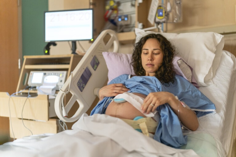 A pregnant woman lies in a hospital bed, wearing a blue hospital gown. Medical monitors and equipment are visible beside her as she rests her hands on her belly, with a fetal monitor attached. The room, which appears to be a maternity ward, is prepared for potential labor induction methods.