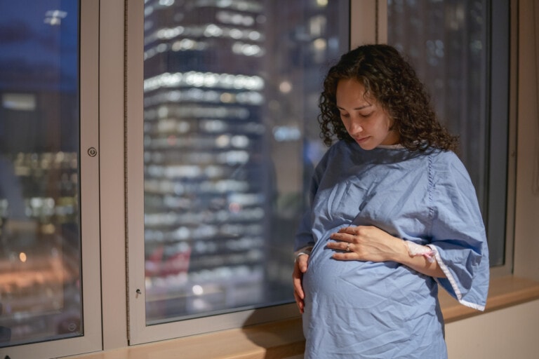 A pregnant woman wearing a hospital gown stands by a window, looking down and gently holding her belly. The window shows the blurred lights of a cityscape at night, as she contemplates not just the future but also the cost of having a baby.