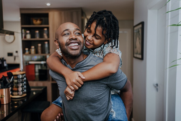 A man is carrying a child on his back, both of them smiling amidst the hustle and bustle of their modern kitchen with wooden cabinets and various items on the countertops. Despite the hidden struggles of parenting, their joy is evident as the child wraps their arms around his shoulders.