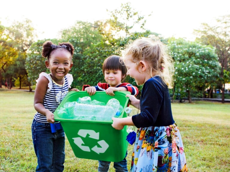 Three children are outdoors, smiling behind a green recycling bin filled with plastic bottles. They are standing on grass with trees and foliage in the background, showcasing one of the easiest volunteer ideas for kids—recycling.