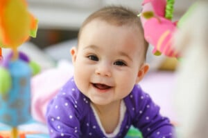A baby girl, possibly named one of those unique girl names starting with H, is wearing a purple onesie with white polka dots and lying on her stomach, smiling. She is surrounded by colorful toys in the slightly blurred background.