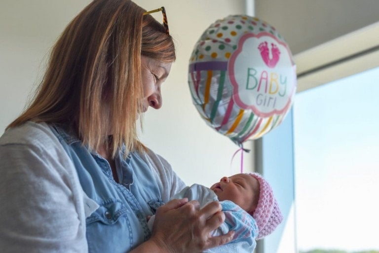 A woman with shoulder-length hair and eyeglasses resting on her head smiles down at a newborn she is holding. A balloon that reads "BABY Girl" is visible in the background. A window with natural light is to the right, highlighting the perfect scene for considering thoughtful gifts to bring a new family.