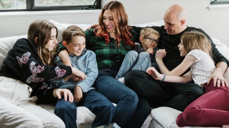 A family of six sits closely together on a couch, embodying the journey from chaos to calm. Two adults and four children, dressed casually, appear happy and relaxed. One child points at another while they all share smiles and comfortable interactions. The background includes a window and the couch.