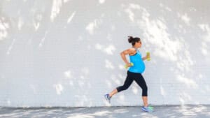 A pregnant woman in a blue tank top and black leggings is running while pregnant, holding yellow dumbbells. She moves gracefully past a white brick wall, with dappled sunlight filtering through the leaves, embodying exercise for pregnant women.