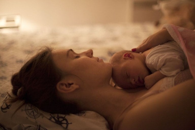 A woman recovering from childbirth is lying on a bed with her eyes closed, holding a sleeping baby on her chest. The baby is swaddled in a light-colored blanket and resting peacefully. The scene appears calm and serene, with soft lighting in the room.