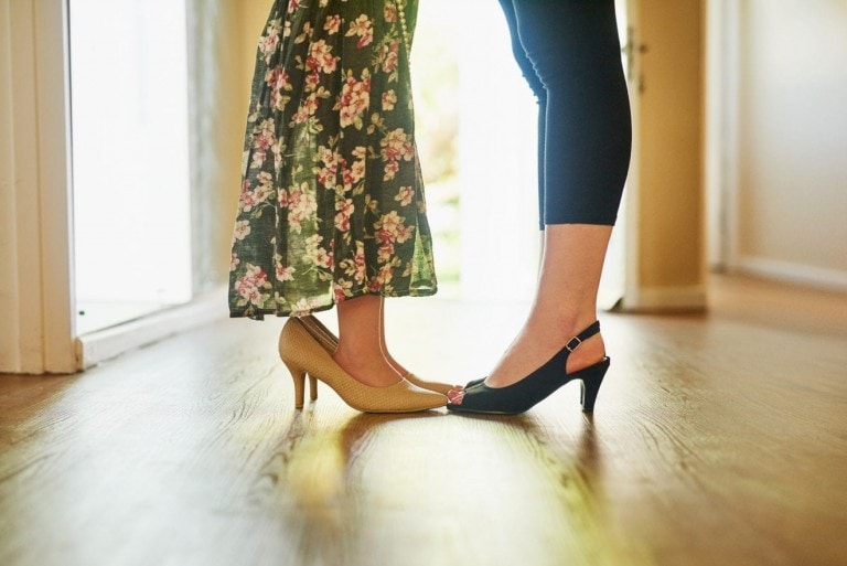 A close-up of two people standing modestly close together indoors, focusing on their legs and feet. One person is wearing a floral dress and beige high heels, while the other is in black pants and black heels. Their feet are nearly touching.