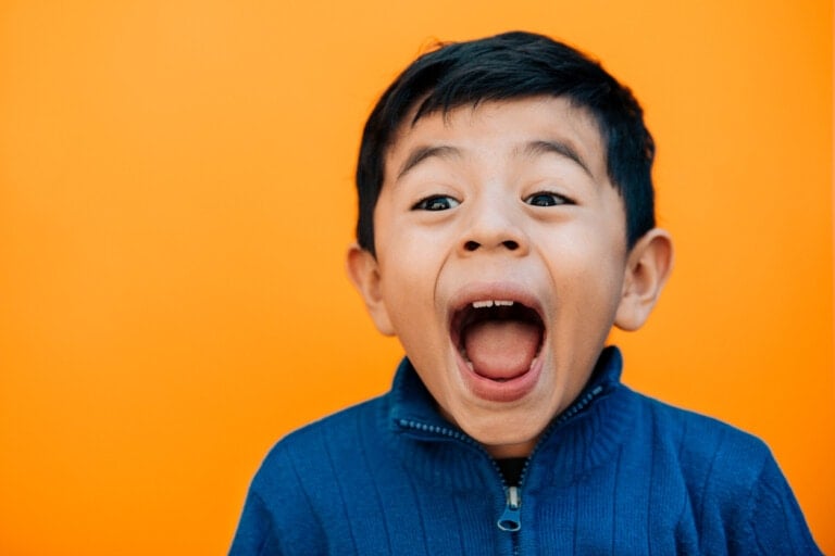 A child with short dark hair and a blue sweater is pictured against an orange background. His mouth is wide open, and his eyes are wide, giving an impression of excitement or surprise—perhaps he just heard some funny jokes for kids!
