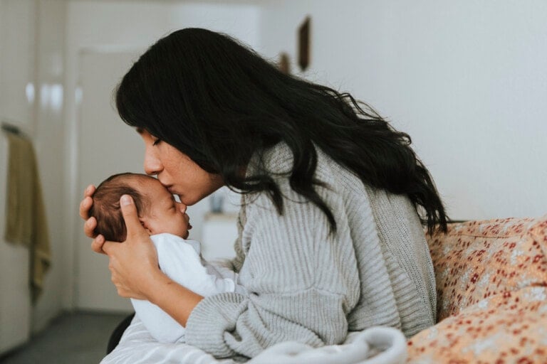 A woman with long dark hair is sitting on a patterned couch, holding a baby close to her chest. She is gently kissing the baby's head. The background shows a doorway and a hanging garment, evoking the tender moments often captured by Becky Vieira.