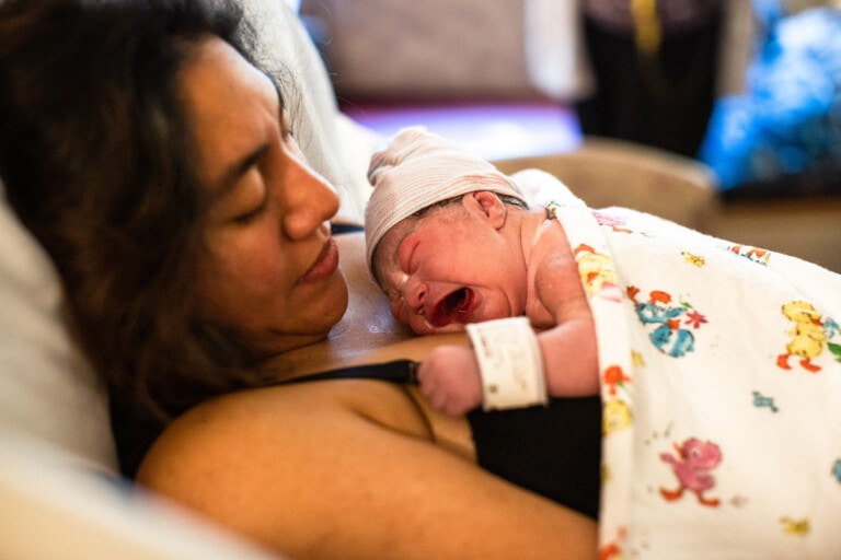 A newborn baby wrapped in a patterned blanket is lying on the chest of a person resting in a hospital bed after a vacuum birth. The baby wears a hat and an ID band while crying. The person, with long hair, appears to be resting.