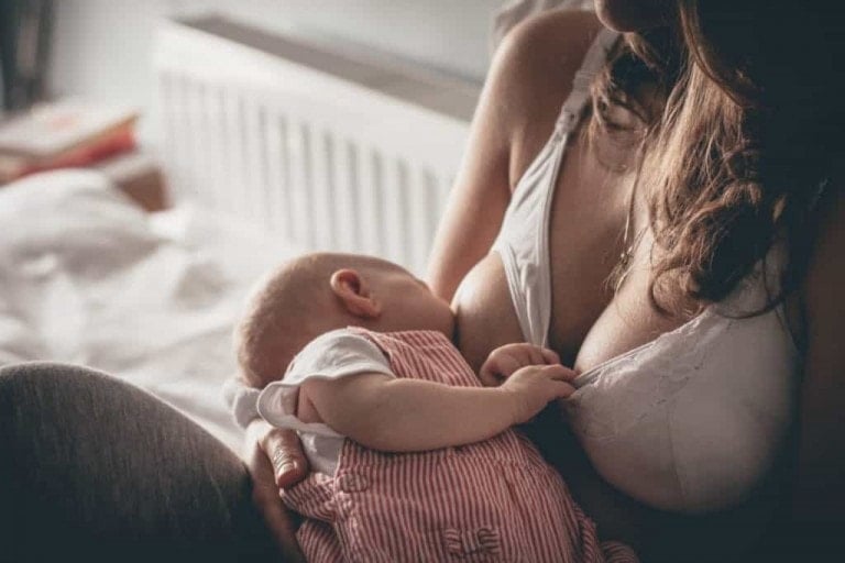 Woman breastfeeding an infant in a dimly lit room. The baby, wearing a red and white striped outfit, is held in the woman’s arms as they sit on a bed. The woman, who underwent breast augmentation before the baby, is dressed in a white bra with her dark hair falling over her shoulder.