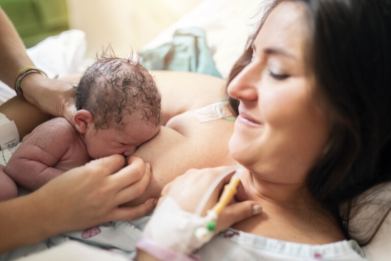 A newborn baby with wet hair is pressed against its mother's chest, breastfeeding for the first time after a c-section. The mother, wearing a hospital gown and with an IV in her hand, is lying on a bed, smiling gently and looking at the baby.