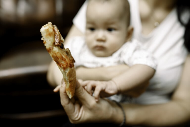 A baby, held by a person in a white shirt, looks intently at a slice of pizza with toppings, which is being held close to it by another hand. The baby appears curious and reaches out toward the pizza, showing an early interest in first foods. The background is blurred.