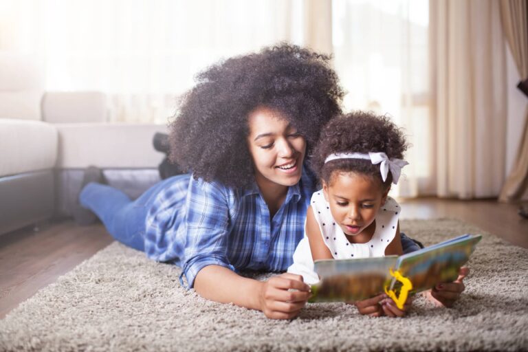 An adult and a young child are lying on a carpeted floor, demonstrating how to teach a child to read. The adult, wearing a blue plaid shirt, smiles while the child, dressed in a white polka-dot dress with a bow, is intently looking at the book. A couch is visible in the background.