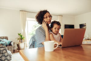 A working mom is sitting at a wooden table, holding a phone to her ear with one hand while a young child is on her lap using a laptop. A white mug is placed on the table in the foreground, capturing the essence of her daily routine. The room is well-lit with a large window in the background.