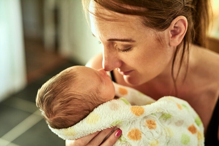 A woman gently holds a swaddled baby close to her face, her nose touching the baby's forehead. The baby, wrapped in a soft blanket with a polka dot pattern, appears to be asleep. The serene moment draws focus on them, evoking the tenderness of a mother named after the Gaelic word for wolf.