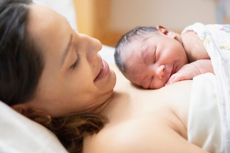 A woman is lying in bed, smiling down at a newborn baby who is sleeping on her chest. The baby, wrapped in a blanket, appears to be resting comfortably. The background is out of focus, placing emphasis on the woman and the baby. Listening to a VBAC podcast nearby adds to their serene moment.