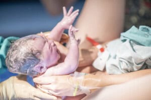 A newborn baby is being pulled up and held immediately after birth. The baby has a wet head and is crying, with eyes closed. Gloved hands support the baby, with a medical bracelet visible on the adults wrist. The setting is likely a hospital or birthing center.