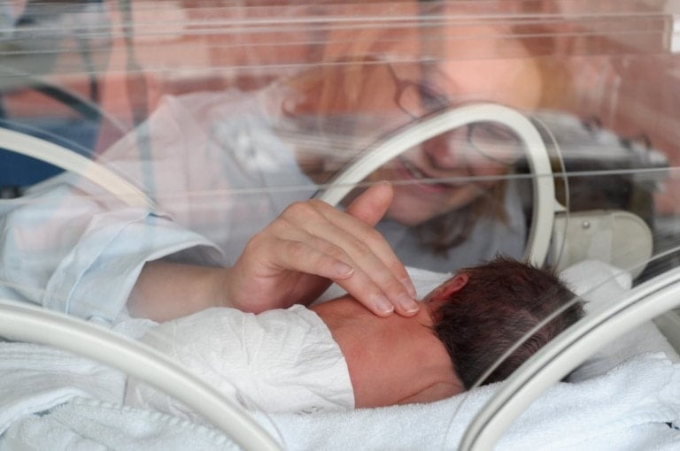 A newborn baby lies in an incubator, covered with a white blanket. A NICU mom's hand gently touches the baby's back through the incubator's access ports. The mom is visible through the clear walls of the incubator, smiling down at her NICU baby.