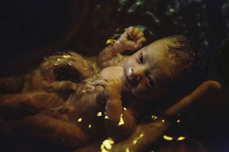 A newborn baby is held in dimly-lit water, with golden reflections on the surface. The baby has wet hair and looks up with eyes partially open, evoking a serene moment synonymous with the gentle experience of an orgasmic birth. The infant appears to be in the hands of an adult, partially submerged.