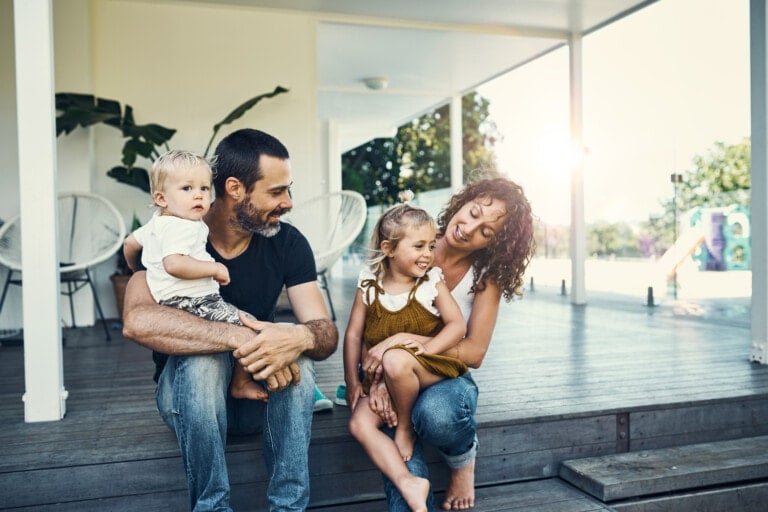 A family of four sits on the steps of a porch. A man holds a toddler while a woman with curly hair, embodying what it means to be a good wife, holds a young girl. The mother and daughter are laughing together. The porch has plants and outdoor chairs, with sunlight streaming in from the right.