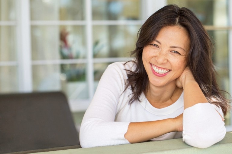Smiling woman with medium-length dark hair, wearing a white long-sleeve top, leaning on a surface with her right elbow and resting her head on her right hand. The serene mood is perfectly captured against the background of large white-framed glass windows.