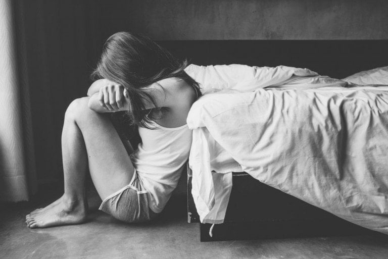 A person with long hair sits on the floor beside a bed, partially leaning on it with their knees drawn up and head resting on their arms. The room is dimly lit, creating a somber atmosphere; a moment of quiet reflection following pregnancy and infant loss. The person is dressed in a light shirt and shorts.