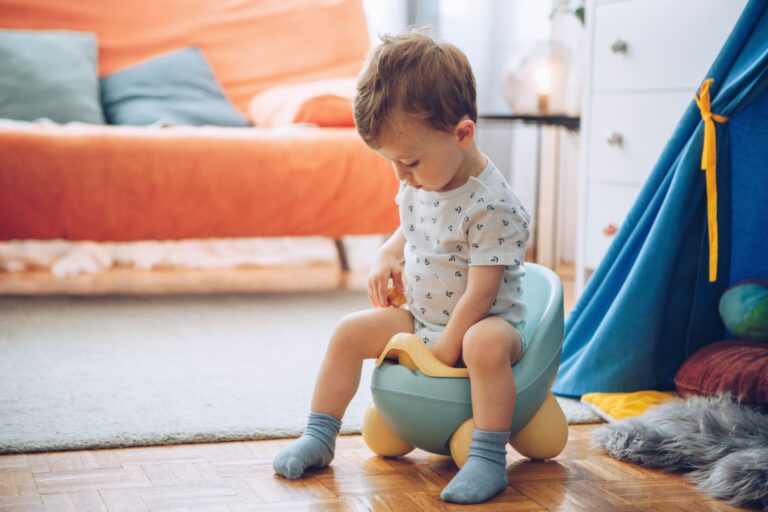A toddler is seated on a potty in a living room with a wooden floor. The child is wearing a patterned shirt and blue socks. There is an orange sofa and cushions in the background, along with a blue play tent and children's toys on the floor—a scene that dispels many myths about potty training complexities.