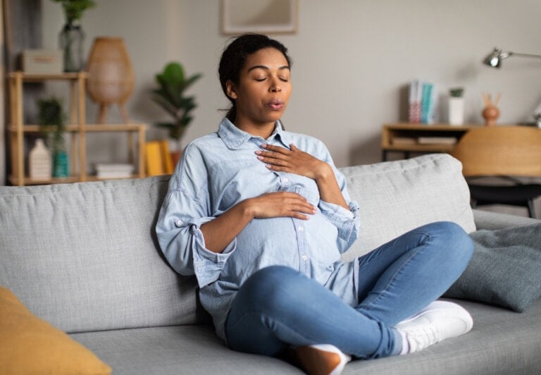 A pregnant woman sits cross-legged on a gray sofa, wearing a denim shirt and jeans. Her eyes are closed, one hand on her chest and the other on her belly, taking deep breaths as pregnancy hormones run through her body. The room has modern decor with shelves and plants in the background.