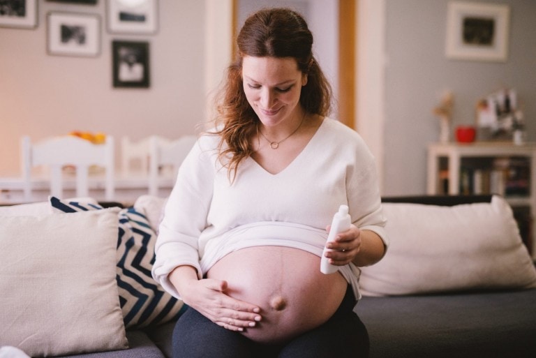 A pregnant woman sits on a couch, looking down and smiling while applying lotion to her bare belly, embracing the changes like pregnancy stretch marks. She wears a white top, and the cozy living room behind her includes a couch, pillows, framed pictures on the wall, and a table with various items.