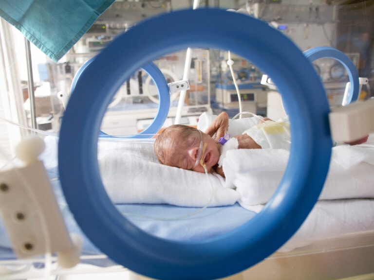 A premature baby lies in an incubator in a neonatal intensive care unit. The baby is connected to medical tubes and monitors, and is wrapped in white blankets. The image, possibly reflecting concerns about the U.S. Premature Birth Rate, is seen through a circular opening, likely part of the equipment.