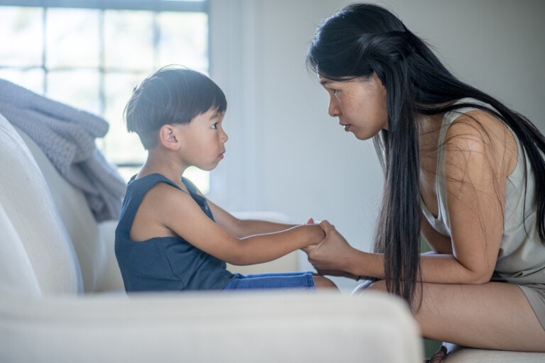 A woman with long hair kneels in front of a young boy seated on a couch. They are holding hands and looking into each other's eyes, engaged in tough conversations. The background features a window with a light curtain partially drawn aside.