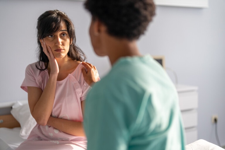 A woman sits on a bed, wearing a pink outfit, her facial expression one of concern as she rests her hand on her cheek. She is looking at another person in a teal outfit who is facing away from the camera, seemingly discussing news about a blighted ovum.