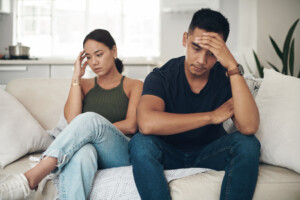 A woman and a man sitting on a couch in a living room, both appearing frustrated and distressed. The woman is looking down with her hand on her head, while the man is looking down with his hand on his forehead; it seems one partner wants a baby, and the other is hesitant.