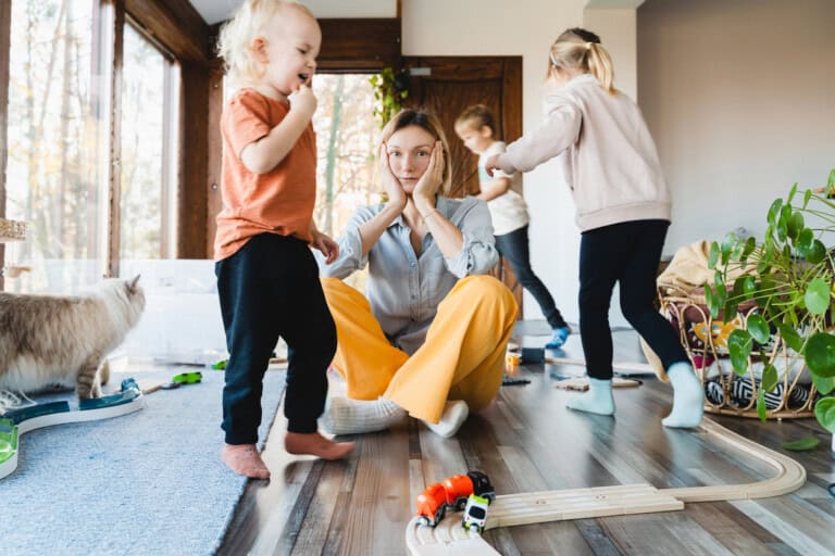 A 21st century mom sits on the floor, hands on her cheeks, looking contemplative. Around her, three children joyfully play with toy trains and tracks. A cat ambles by in the room filled with scattered toys and plants, lit by the light from large windows.