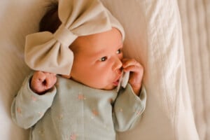 A baby is lying down, wearing a light green outfit with small floral prints. The baby has a large beige bow on their head and is holding a pacifier in their mouth. The background consists of a textured light-colored fabric, perfect for announcing modern girl names.