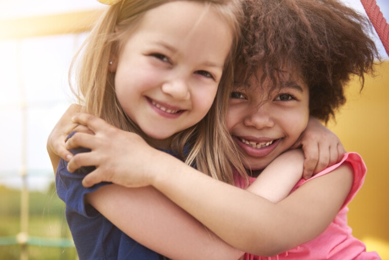 Two children smile at the camera while embracing each other, showcasing the power of words in their radiant expressions. One child has straight, blonde hair and wears a blue shirt, while the other child has curly, brown hair and wears a pink shirt. The background is bright and sunny with outdoor play equipment visible.