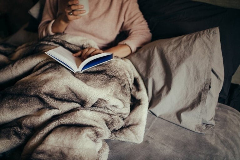 Person wrapped in a soft, fur-like blanket, sitting on a bed with gray sheets and pillows, holding an open book for new parents in one hand. The other hand holds a mug close to their chest. They are wearing a light pink sweater.